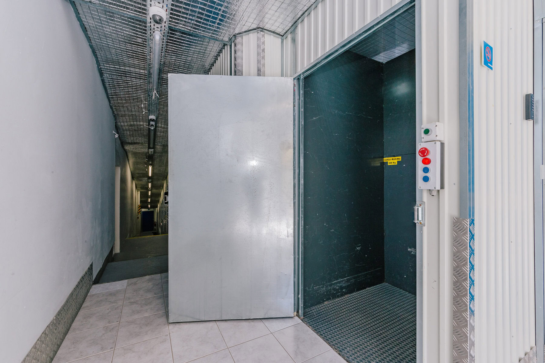 Um elevador industrial espaçoso com uma porta de metal aberta, revelando um interior revestido com paredes e piso de metal texturizado. O painel de controle apresenta botões azuis e vermelhos. A área ao redor é iluminada por luzes de teto.