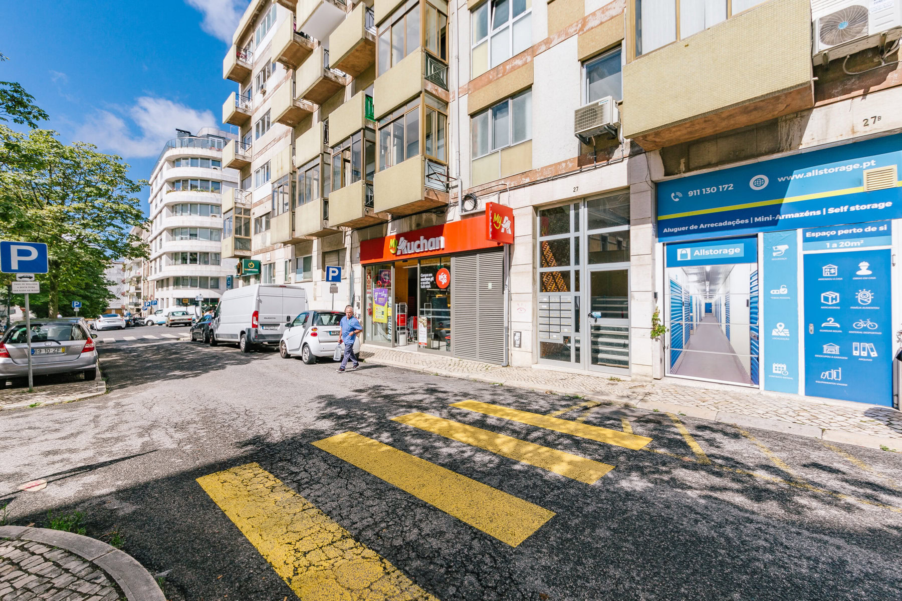 A vista da rua mostra um prédio com vários negócios, incluindo uma pequena loja Auchan. Linhas amarelas de travessia de pedestres estão na estrada, e árvores margeiam a rua. Veículos estão estacionados ao longo da calçada, e um negócio de autoarmazenamento azul é visível.