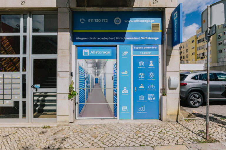 Fachada de um negócio de self-storage com sinalização azul com ícones e texto brancos. A entrada com porta de vidro mostra uma imagem de corredores de armazenamento. Um carro prateado está estacionado nas proximidades, e os edifícios adjacentes são visíveis sob um céu parcialmente nublado.