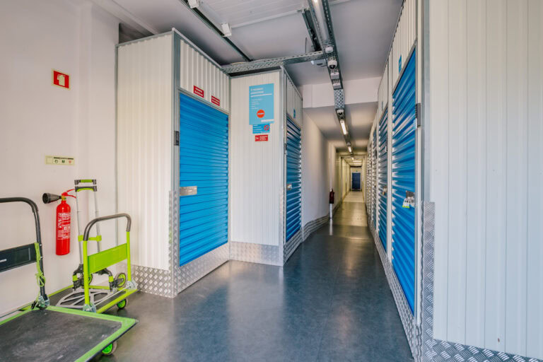 ChatGPT Esta imagem mostra um corredor interno de um armazenamento de self-storage da marca Allstorage Self Storage. Há várias portas de metal azul alinhadas ao longo do corredor, com as portas à direita sendo visíveis mais de perto. Cada porta tem uma pequena tranca de metal. À esquerda do corredor, há dois carrinhos de transporte com estrutura verde e plataforma preta. Há também um extintor de incêndio vermelho montado na parede esquerda. O corredor é bem iluminado com luzes fluorescentes no teto. A sinalização de emergência e instruções estão fixadas nas portas e paredes. O chão é de material cinza escuro, e a parte inferior das paredes é revestida com uma placa metálica antiderrapante.