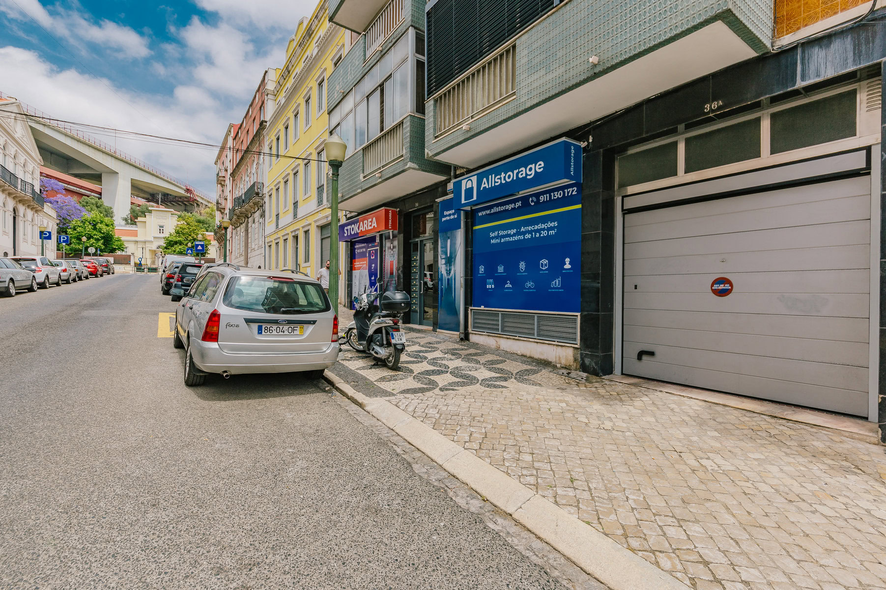 Vista da rua de uma cena urbana estreita com carros estacionados e uma scooter. Um prédio exibe placas para Allstorage e KOPA LIBRA. Uma ponte é visível ao fundo, e há várias plantas ao longo da rua.