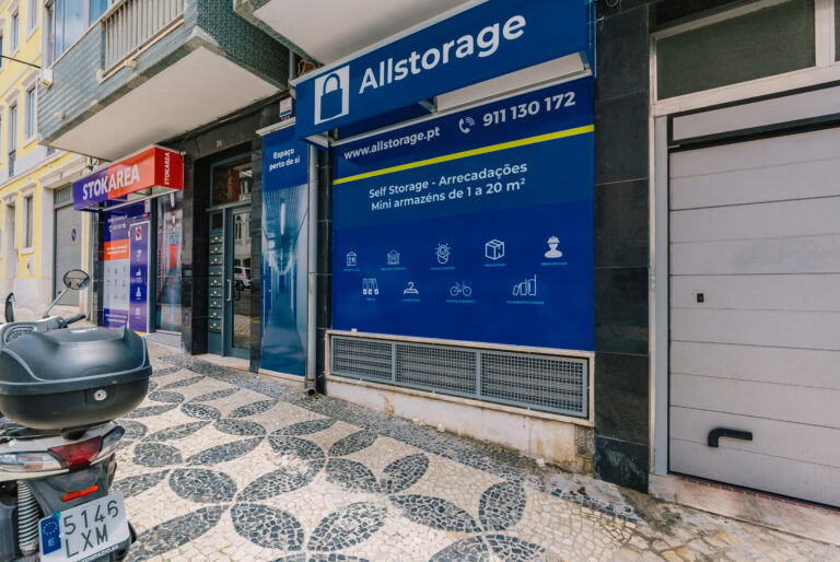 Vista da calçada de um negócio de armazenamento com uma fachada azul exibindo a sinalização Allstorage. O texto inclui detalhes de contato e serviços. Uma motocicleta está estacionada nas proximidades, e outro negócio de armazenamento é visível ao fundo.