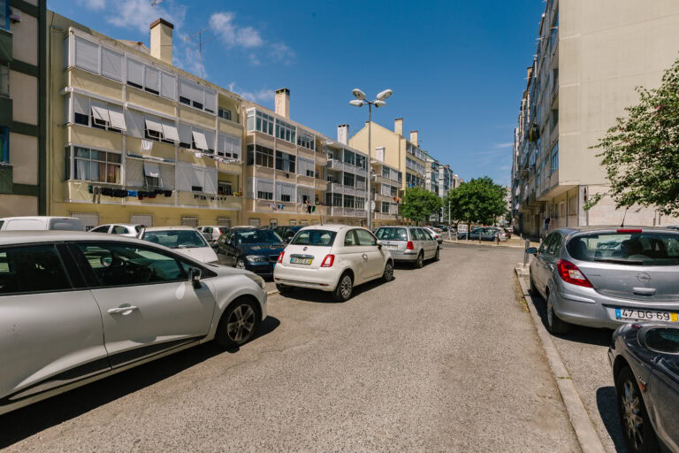 Uma rua urbana tranquila com carros estacionados em ambos os lados. A rua é ladeada por prédios de apartamentos com sacadas. Um céu azul claro é visível com algumas nuvens, e árvores são visíveis no final da rua.