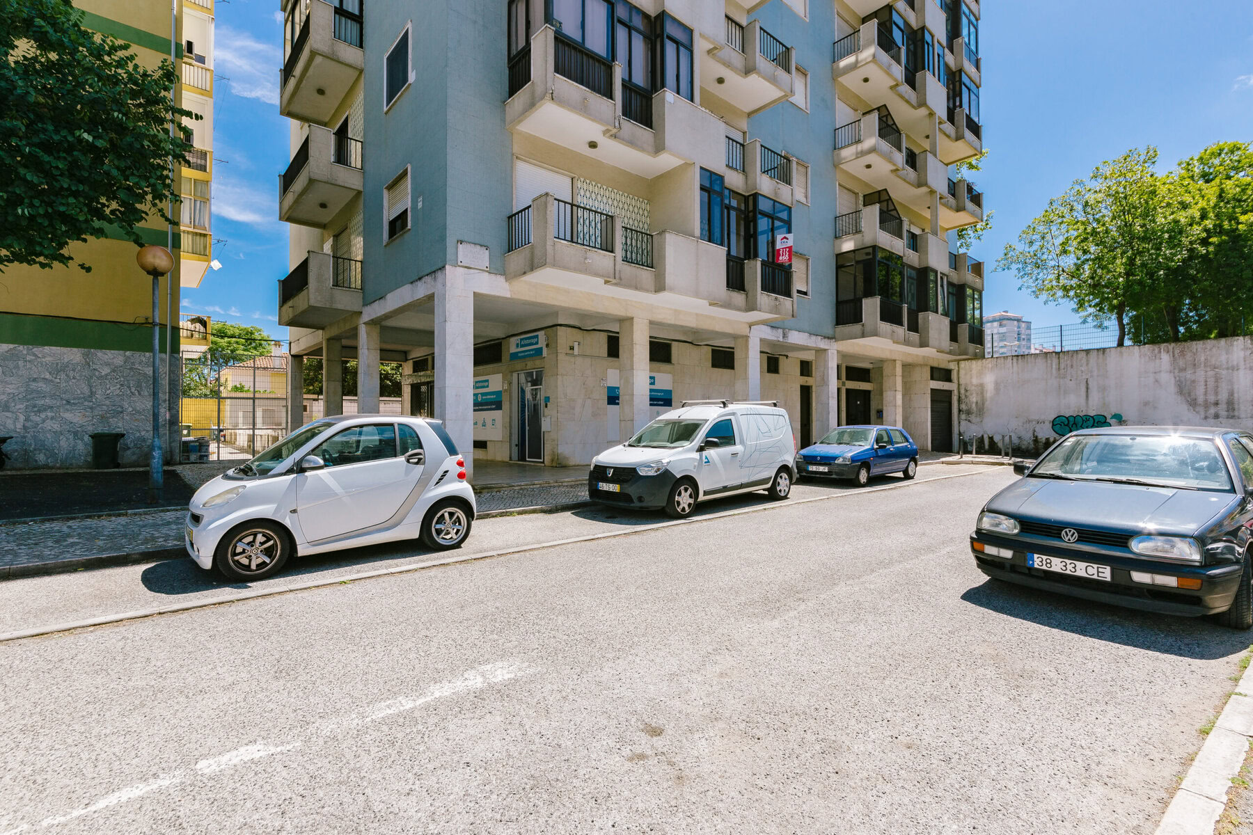 A vista da rua mostra carros estacionados, incluindo um carro smart branco, uma van branca, um sedan azul e um sedan preto, ao lado de um prédio alto de apartamentos com varandas. Uma árvore e um muro de pedra são visíveis ao fundo sob um céu azul claro.