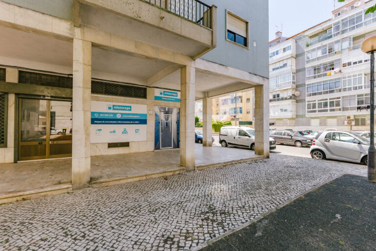 Vista da rua da entrada de um prédio com sinalização azul e branca. Carros estão estacionados ao longo do caminho de paralelepípedos, e outros prédios são visíveis ao fundo. A porta e as janelas refletem um dia claro e ensolarado.