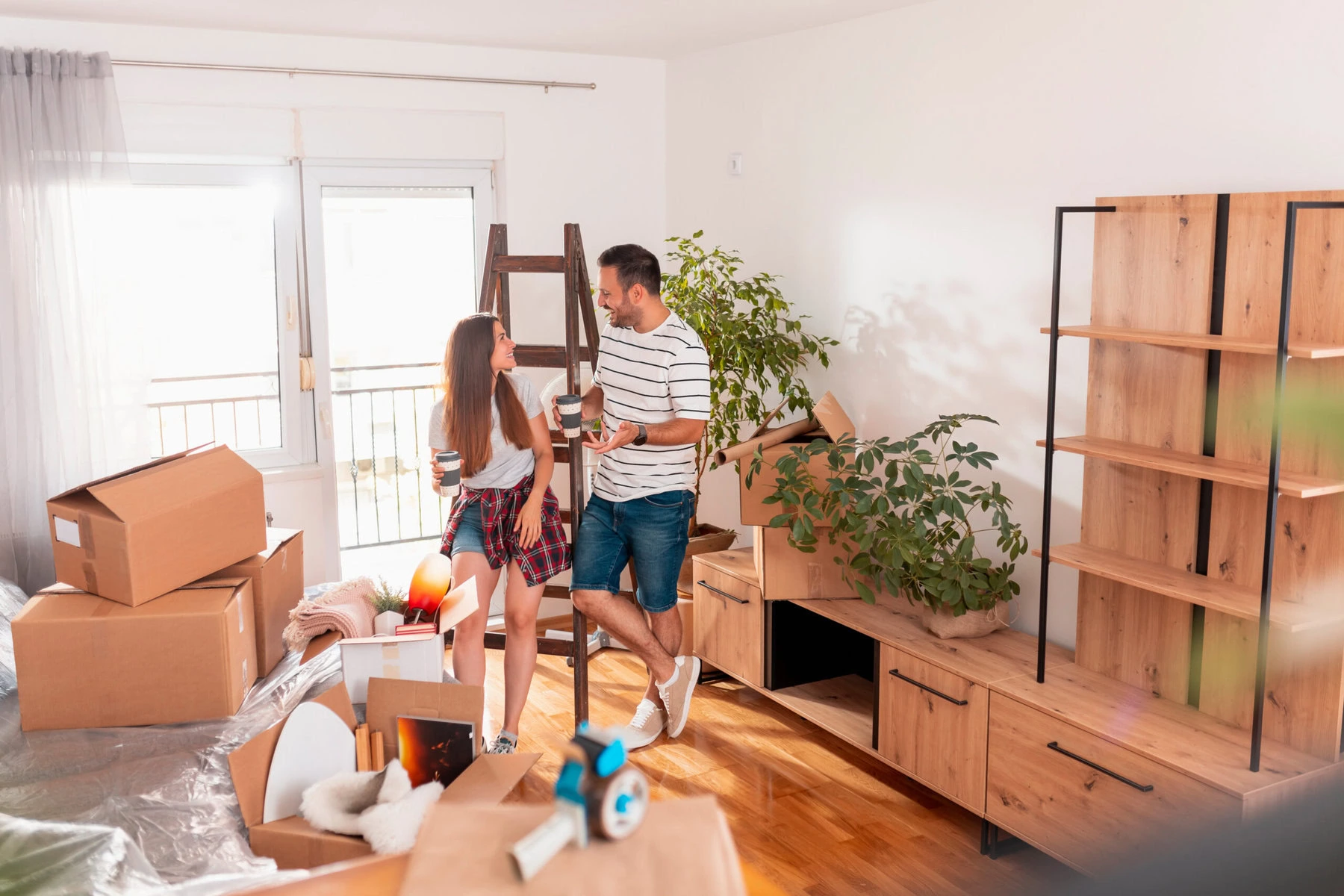 Um casal está em uma sala de estar iluminada, cercado por caixas de mudança desempacotadas e móveis de madeira, abraçando a arte da organização de casa. Segurando xícaras e sorrindo, eles aproveitam a luz do sol que entra por uma janela, destacando uma grande planta ao lado deles.