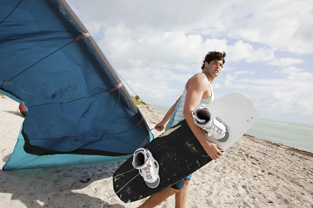 Uma pessoa está caminhando em uma praia arenosa, carregando uma prancha de kiteboard preta e branca e usando um arnês preso a uma grande pipa azul. O céu está parcialmente nublado, e o oceano é visível ao fundo.