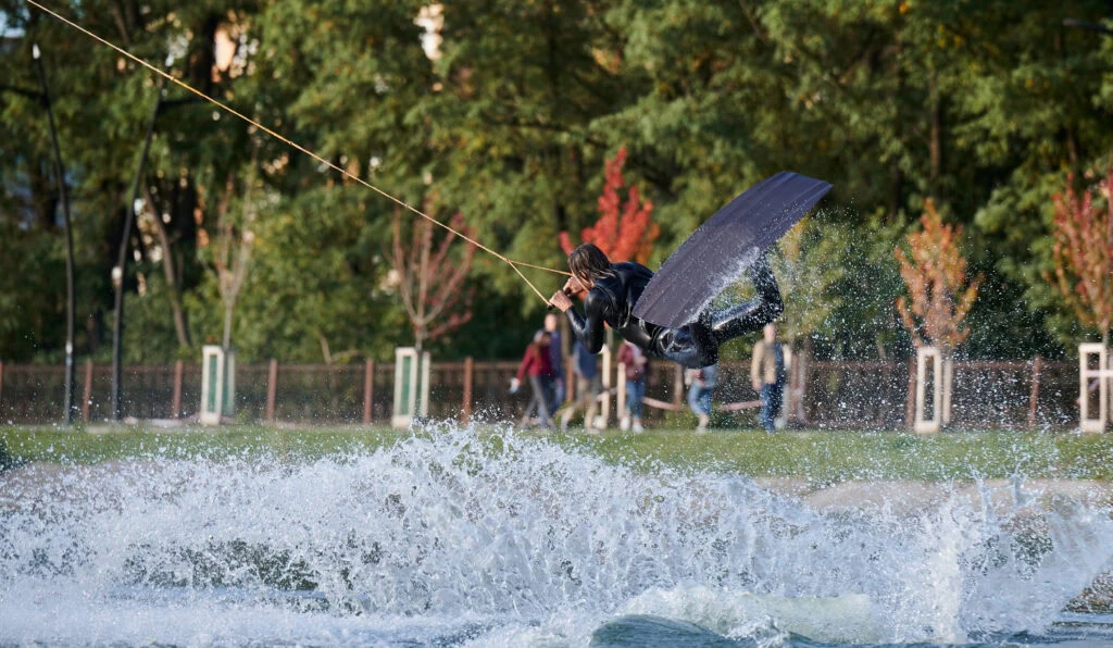 Uma pessoa praticando wakeboard está realizando um salto em um lago, criando um splash. Ela está usando equipamento preto e segurando um cabo, com árvores e algumas pessoas visíveis ao fundo em um dia claro.