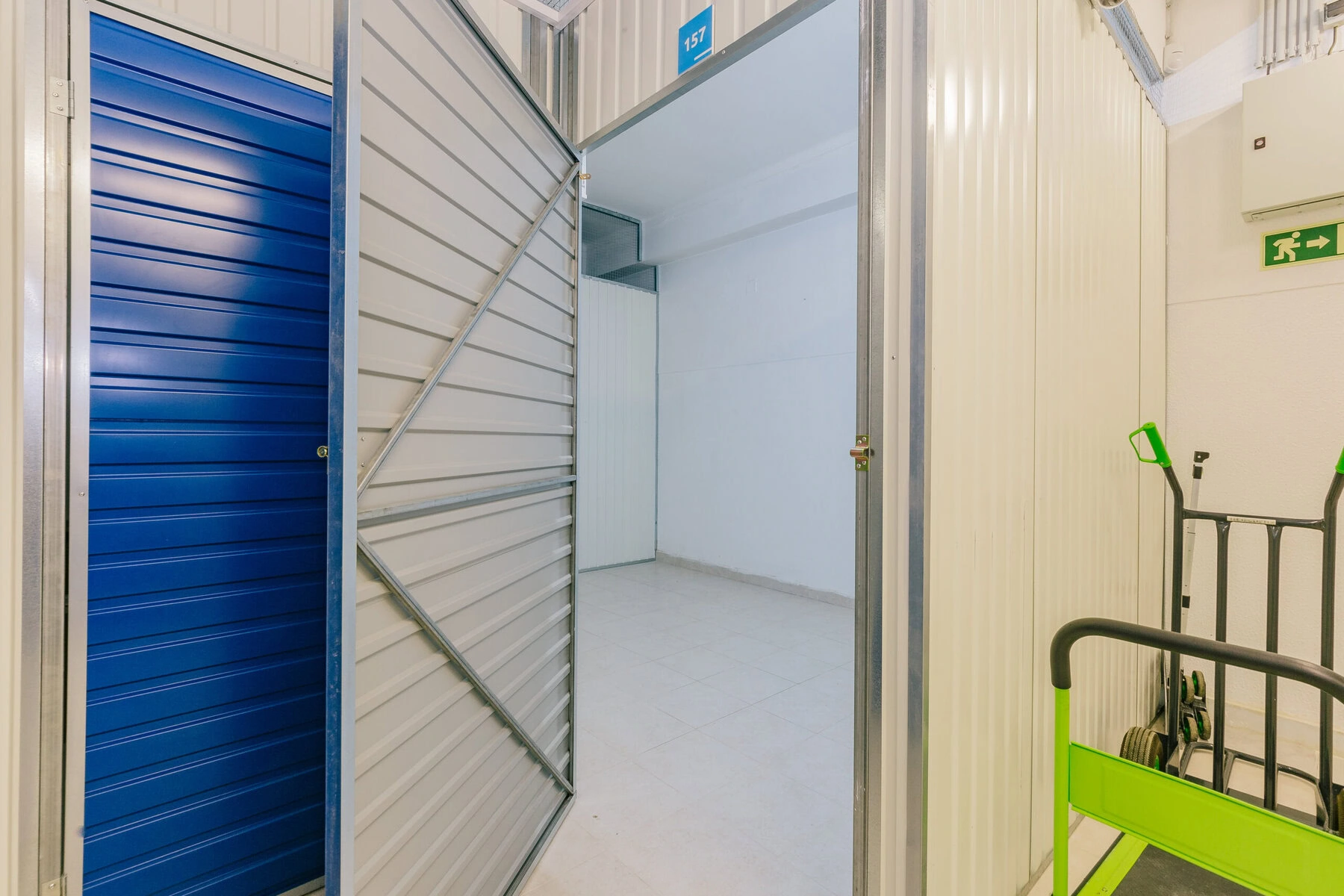 Allstorage Self storage Uma unidade vazia da Allstorage com paredes brancas e piso de cerâmica. O espaço de self storage tem uma porta de metal parcialmente aberta com uma superfície interna azul. À direita, um carrinho de mão verde e preto é visível, e uma placa de saída de emergência está pendurada na parede, típica da nossa localização em Almada. Arrecadacoes e armazens para alugar