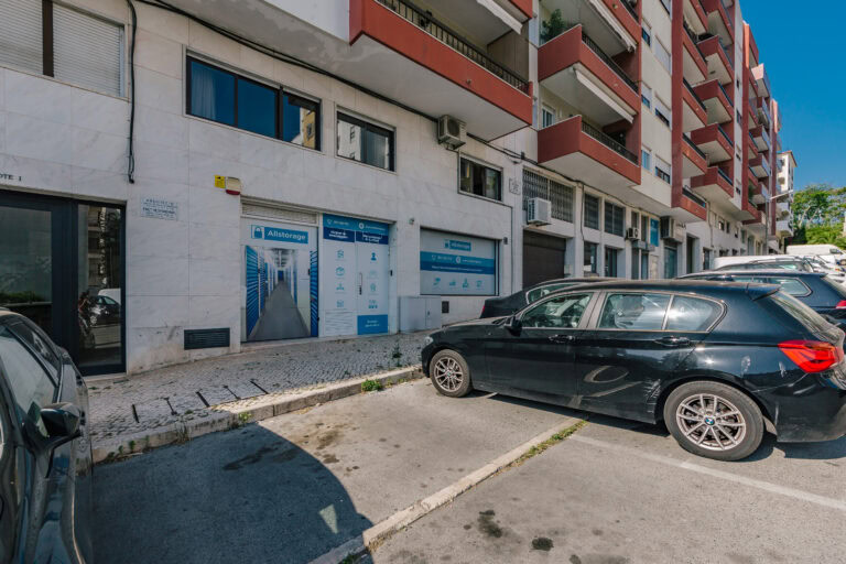 Allstorage Self storage Uma rua da cidade em Sete Rios, Lisboa, exibe um edifício de vários andares com uma fachada de loja chamada Allstorage. Enfeitado com imagens de self-storage, dois carros estão estacionados na frente, emoldurados por um céu azul claro e algumas árvores, capturando a essência do charme urbano português. Arrecadacoes e armazens para alugar