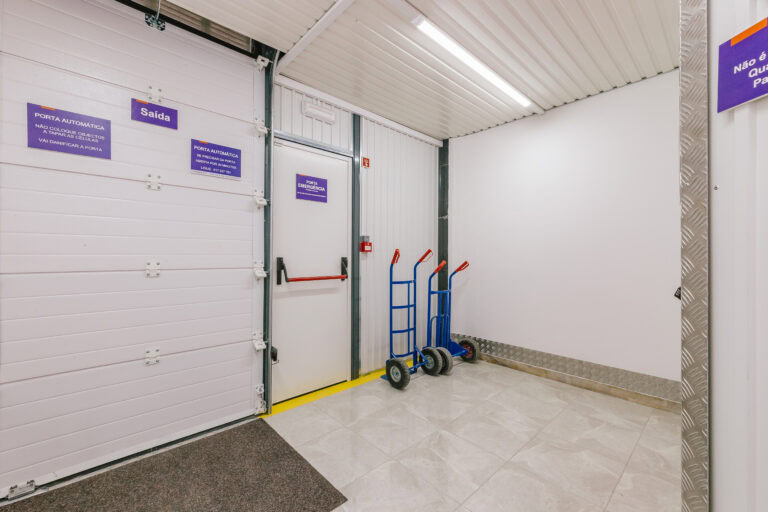 Allstorage Self storage Uma área de armazenamento com uma porta de correr branca à esquerda, marcada Saída (Exit) em português. Dois carrinhos de mão azuis estão à direita, ao lado de uma parede branca. O piso é de ladrilho, e um pequeno tapete cinza está na frente da porta. Arrecadacoes e armazens para alugar