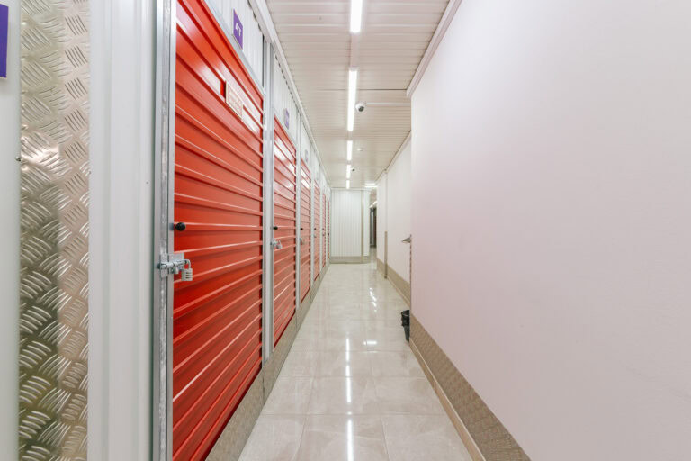 Allstorage Self storage Um corredor em uma unidade de autoarmazenamento com portas de metal vermelhas no lado esquerdo. O piso é feito de ladrilhos brilhantes, e as paredes são pintadas de branco. Luzes fluorescentes no teto iluminam o espaço. Arrecadacoes e armazens para alugar