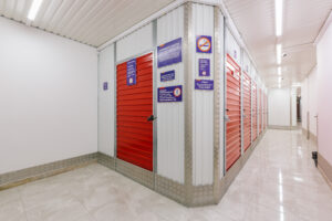 Allstorage Self storage Um corredor bem iluminado com paredes brancas e pisos de ladrilhos brilhantes é alinhado com unidades de armazenamento. Cada unidade tem uma porta vermelha protegida com fechaduras e placas informativas nas paredes. O teto é branco com iluminação de faixa. Arrecadacoes e armazens para alugar