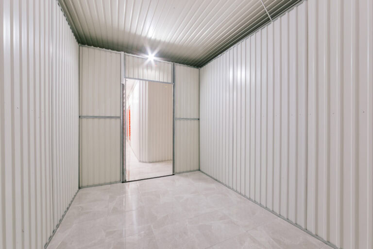 Allstorage Self storage Unidade de armazenamento vazia com paredes e teto de metal corrugado de cor clara. O piso é coberto com ladrilhos cinza claro. A unidade é bem iluminada, com a porta aberta, levando a um corredor com mais unidades de armazenamento visíveis. Arrecadacoes e armazens para alugar