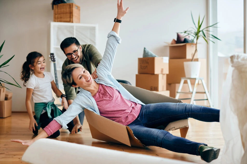 Allstorage Self storage Uma mulher brincalhona senta-se em uma caixa de papelão, com os braços levantados, fingindo deslizar. Ao lado dela, um homem e uma jovem garota riem e observam. A sala está cheia de caixas desempacotadas, indicando uma mudança recente. Arrecadacoes e armazens para alugar