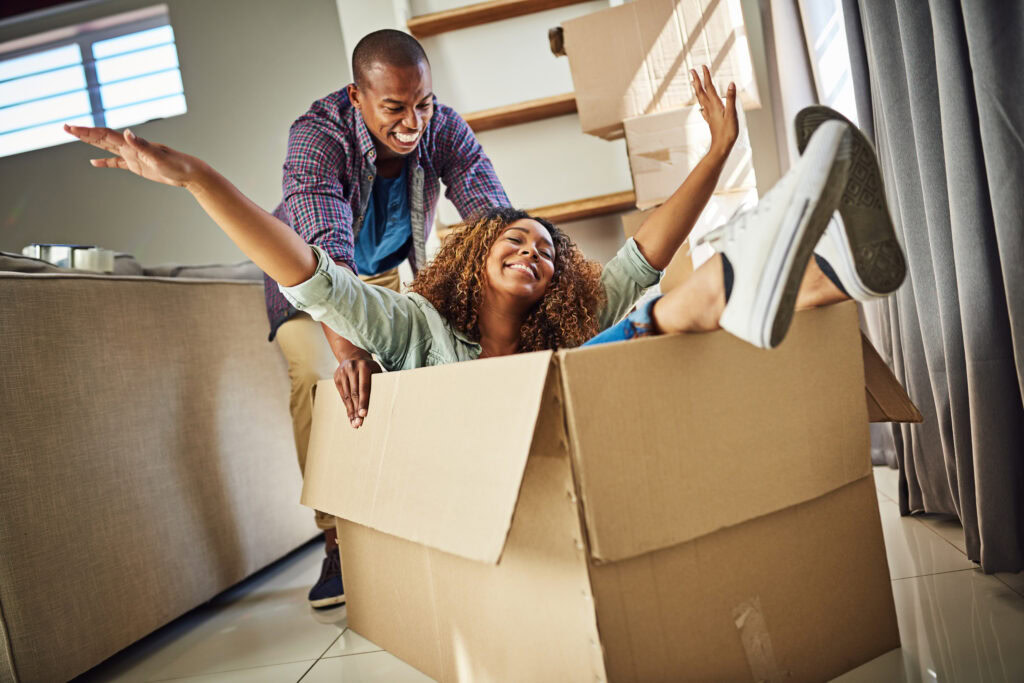 Allstorage Self storage Uma cena alegre de um homem empurrando de brincadeira uma mulher sentada em uma caixa de papelão. Ela tem os braços levantados e sorri largamente, enquanto ele sorri atrás dela. Eles estão dentro de casa, com caixas de mudança empilhadas por perto. Arrecadacoes e armazens para alugar